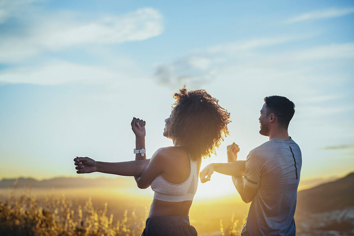 A man and woman stretching in a field with the sun setting in front of them.